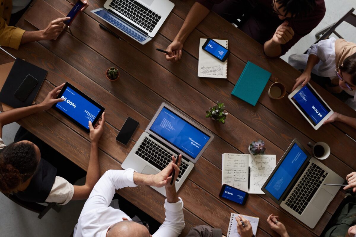 Bovenaanzicht van een vergadering: mensen rond een houten tafel met laptops en tablets met blauwe presentaties, omringd door notities, koffiekopjes en kleine planten.