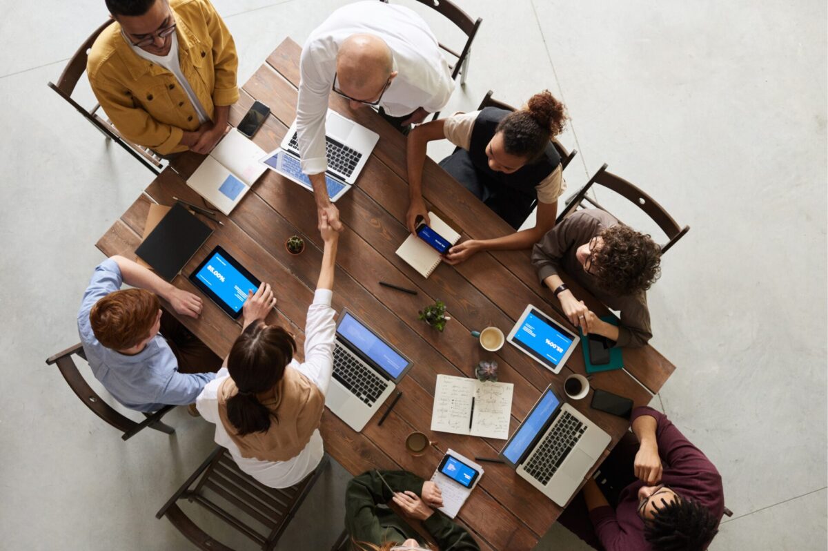 Bovenaanzicht van een team dat rond een houten tafel vergadert met laptops en tablets, terwijl twee collega’s elkaar in het midden de hand schudden.