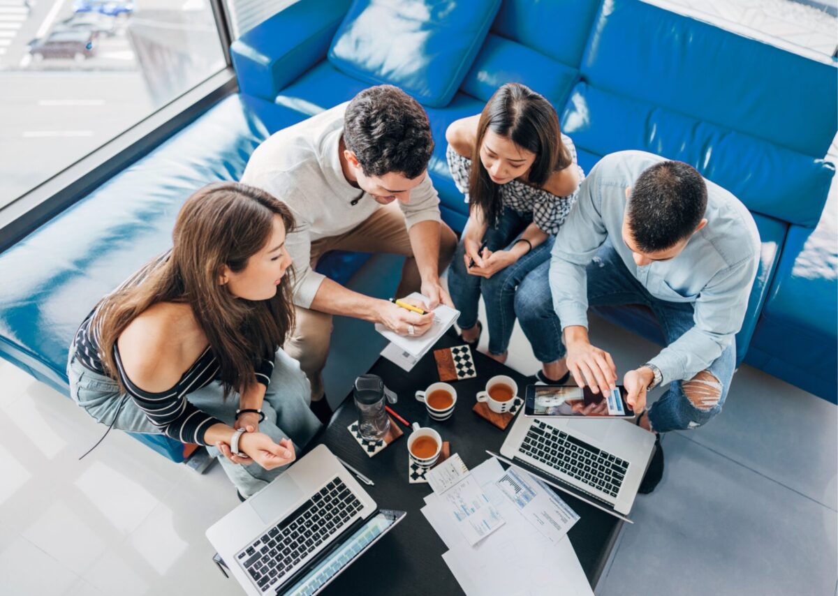 Bovenaanzicht van vier mensen die samenwerken rond een salontafel met laptops, notitieblok en papieren met grafieken, naast kopjes koffie in een informele kantoorruimte met blauwe banken.