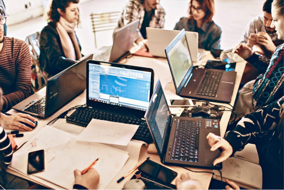 Een groep mensen werkt samen rond een tafel vol laptops, smartphones en papieren schetsen, terwijl op de schermen ontwerpsoftware (CAD) met blauwdrukken te zien is.