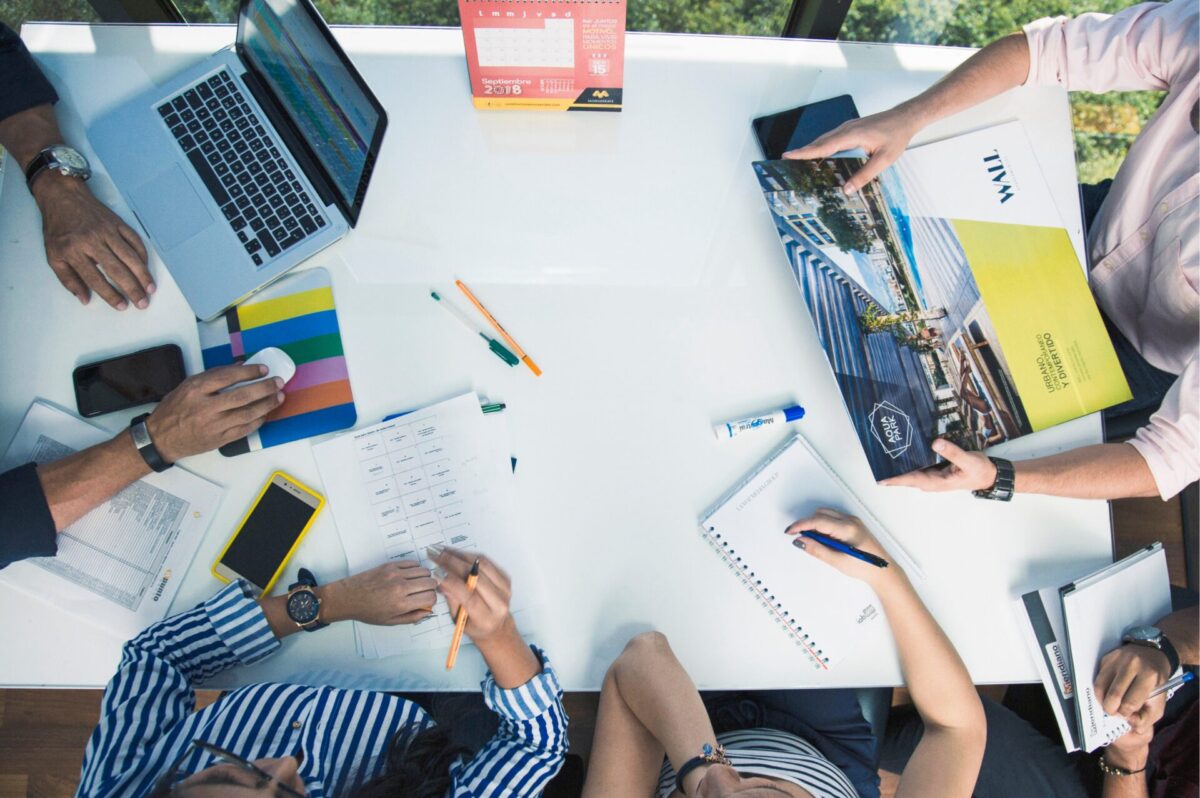 Bovenaanzicht van een teamvergadering rond een witte tafel met laptop, notitieblokken, smartphones, pennen en een kleurrijke brochure terwijl deelnemers notities maken en plannen bespreken.