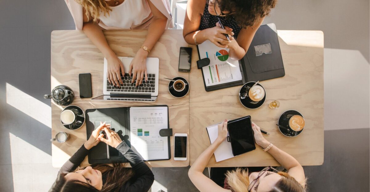 Bovenaanzicht van vier mensen die samenwerken aan een houten tafel met laptop, tablet, smartphones en documenten met grafieken, naast koffiekopjes en een theepot.