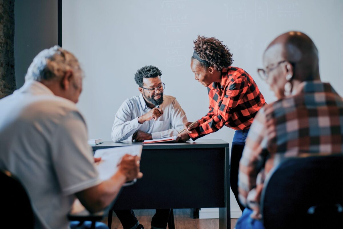 Een docent helpt een student aan een bureau tijdens een les, met twee andere cursisten onscherp op de voorgrond en een whiteboard met wiskundige formules op de achtergrond.
