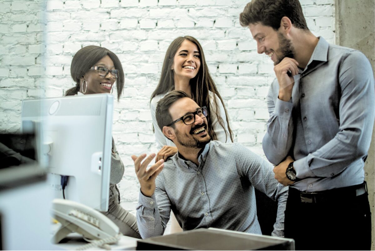 Vier collega’s lachen en overleggen rond een bureau met een computerscherm, voor een witte bakstenen muur in een modern kantoor.