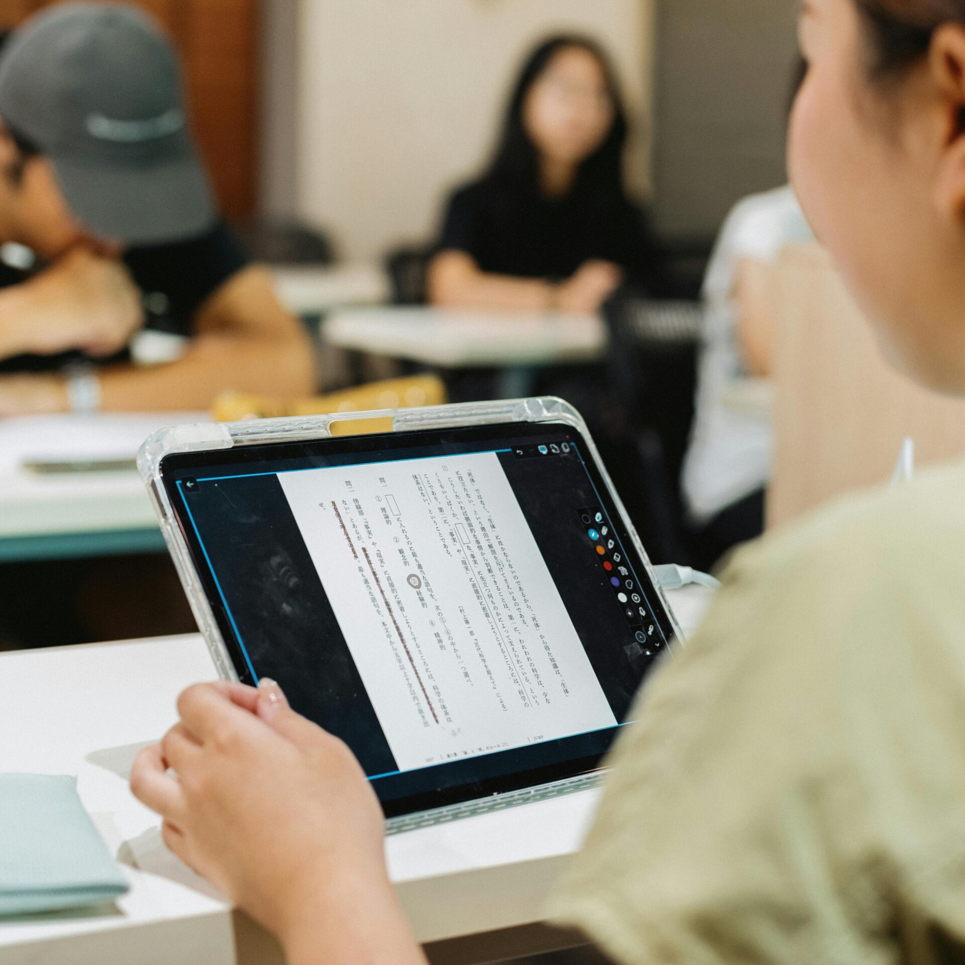 Een student bekijkt lesmateriaal op een tablet in een klaslokaal, met medestudenten onscherp op de achtergrond.