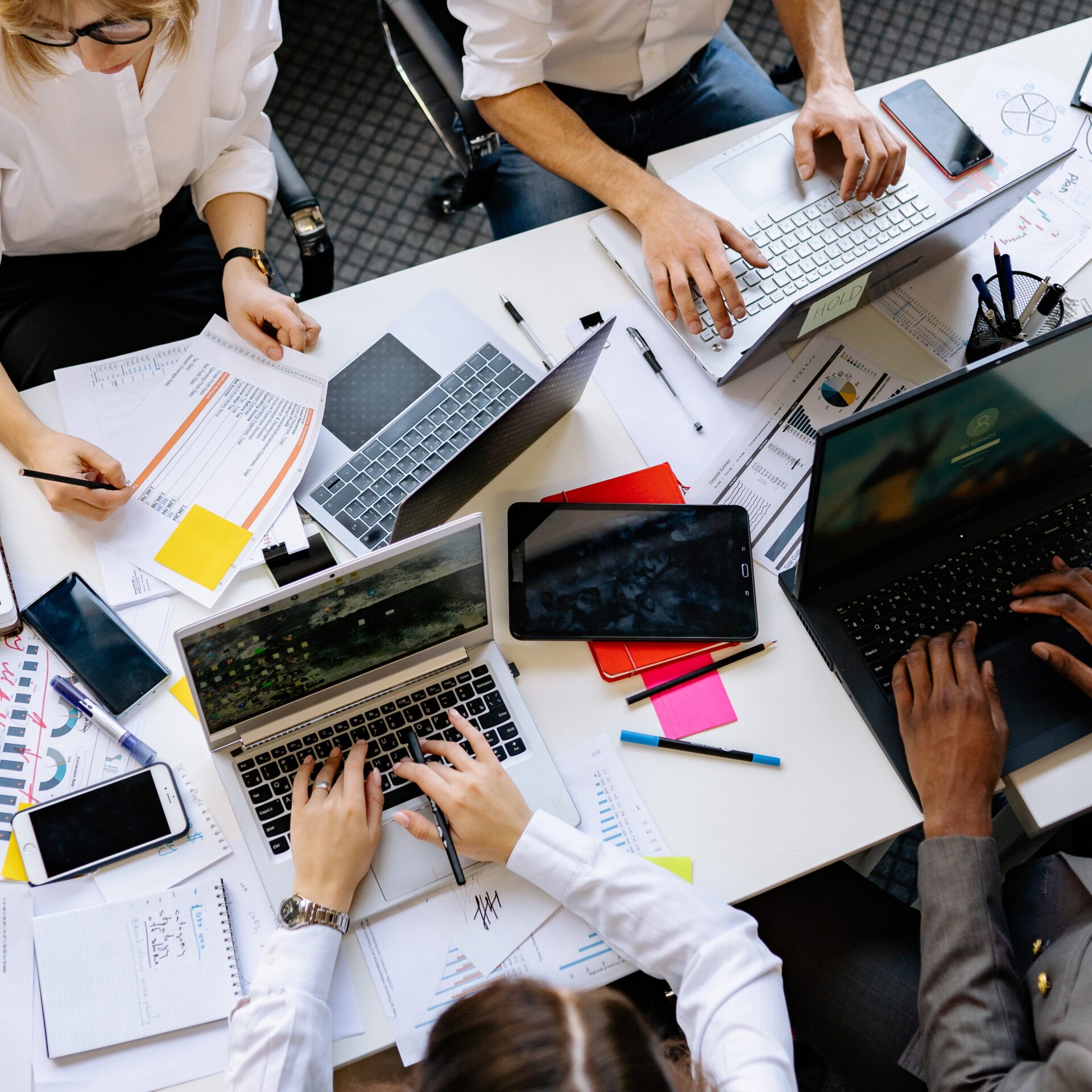Bovenaanzicht van een team dat samenwerkt rond een rommelig bureau met laptops, tablets, smartphones en papieren met grafieken en notities.