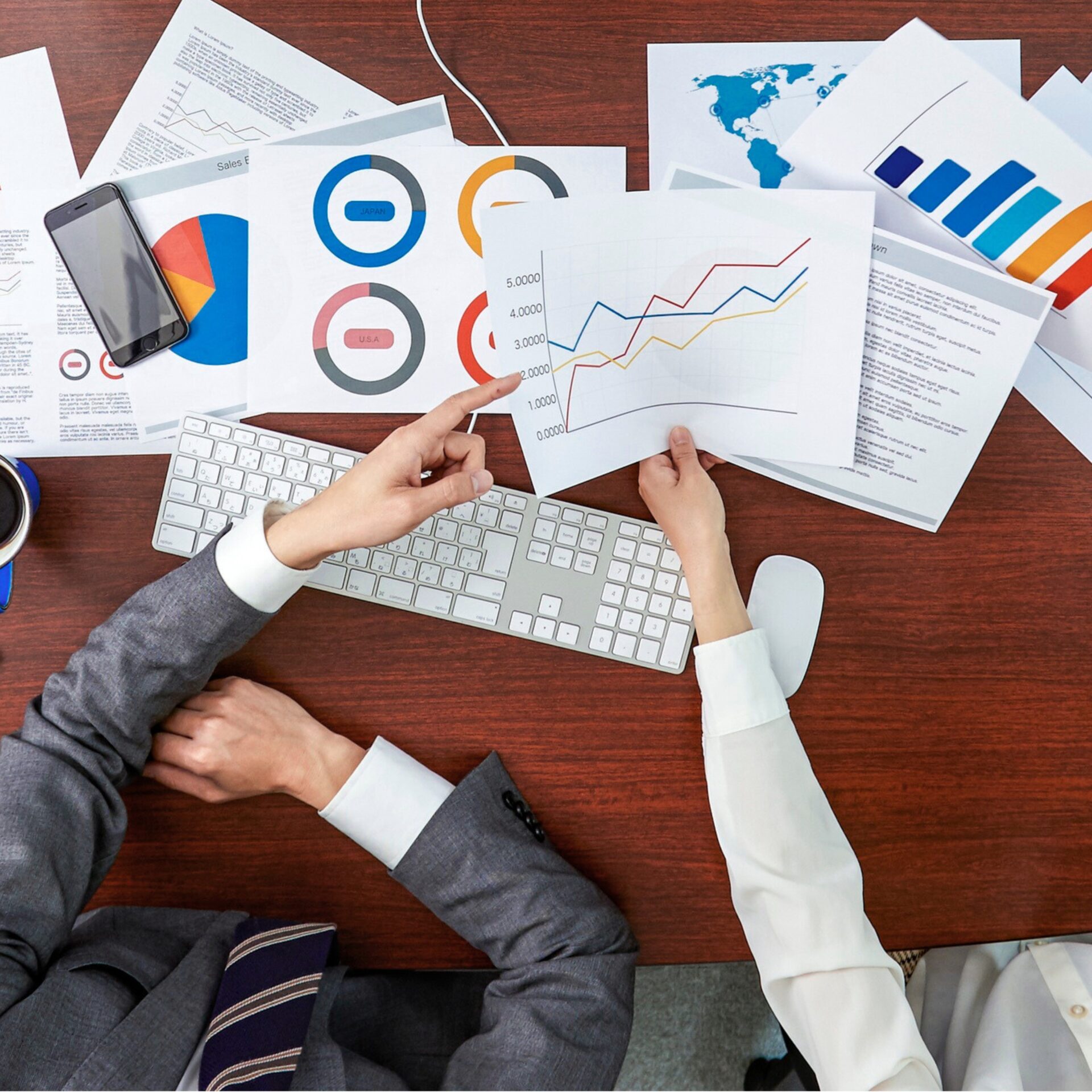 Bovenaanzicht van twee collega’s die aan een bureau financiële grafieken en rapporten bespreken, terwijl één een lijngrafiek met stijgende trend aanwijst tussen geprinte charts, toetsenbord en smartphone.