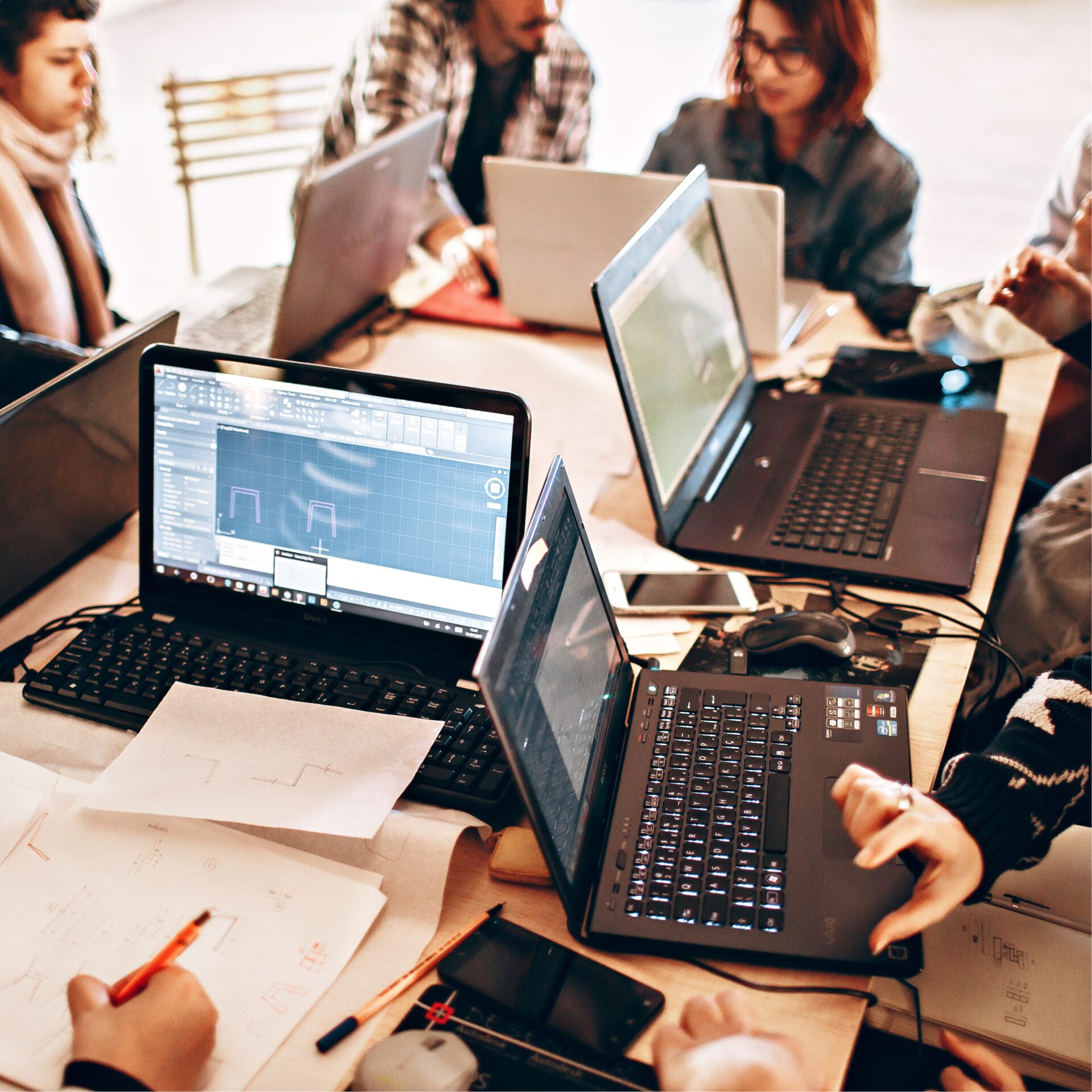 Een groep mensen werkt samen rond een tafel met meerdere laptops, papieren met technische schetsen en een scherm met CAD‑software op de voorgrond.