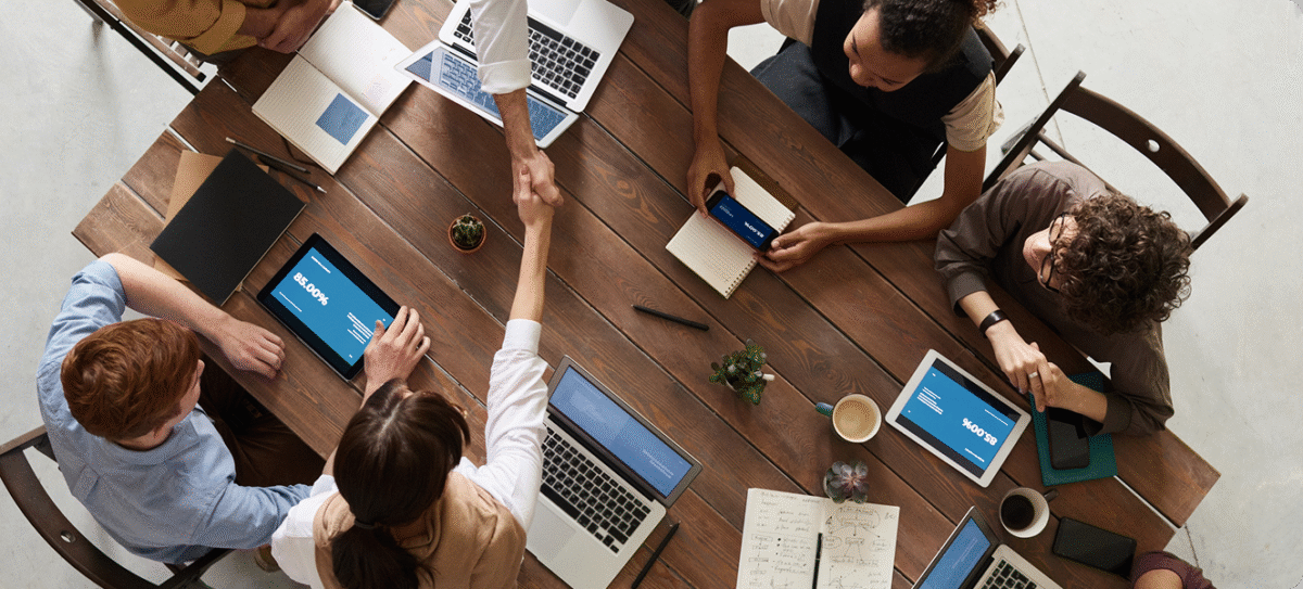 Bovenaanzicht van een teamvergadering rond een houten tafel, met laptops en tablets op tafel terwijl twee collega’s elkaar de hand schudden.