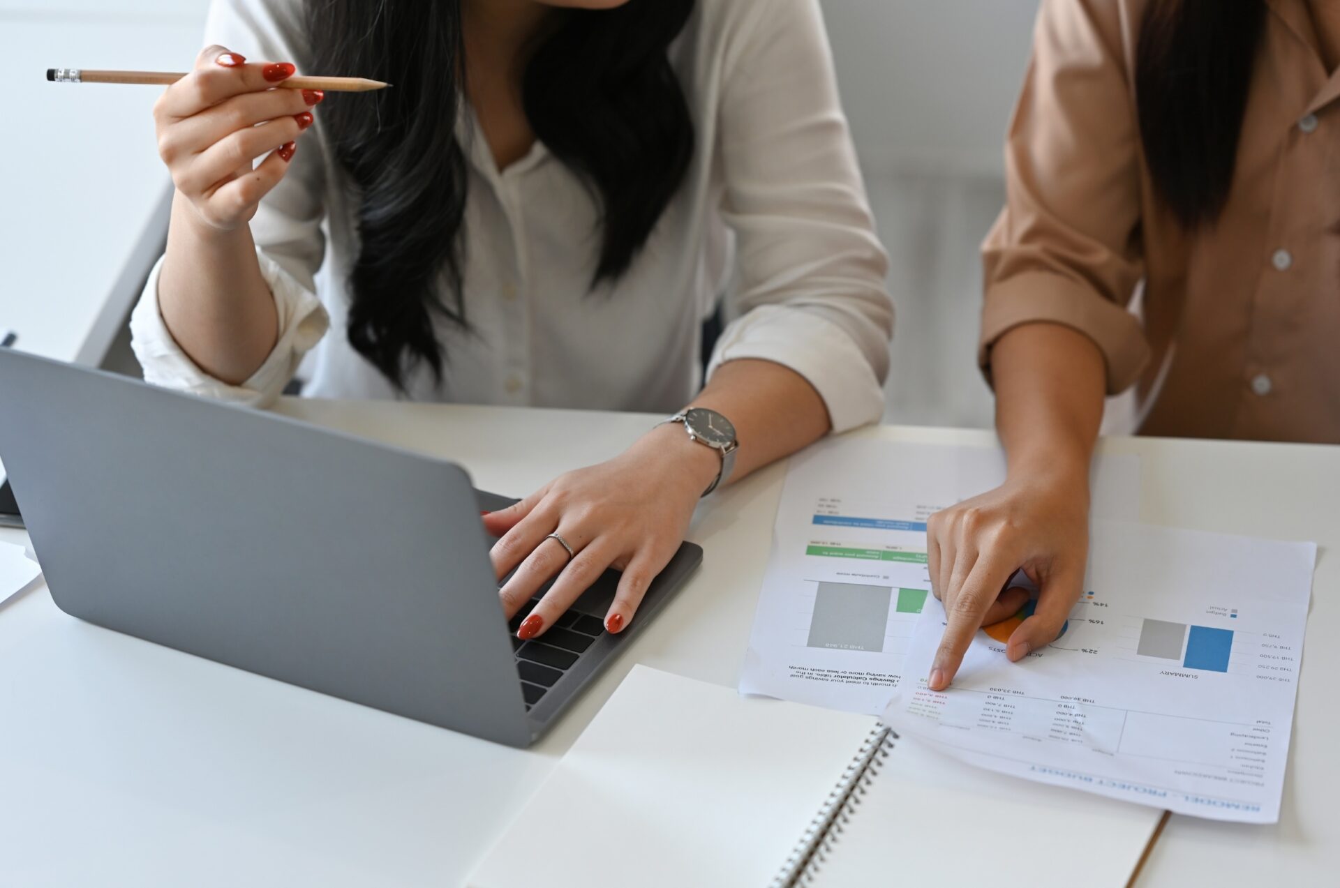 Twee collega’s werken aan een bureau; één typt op een laptop en houdt een potlood vast, terwijl de ander naar grafieken op een geprint rapport wijst.