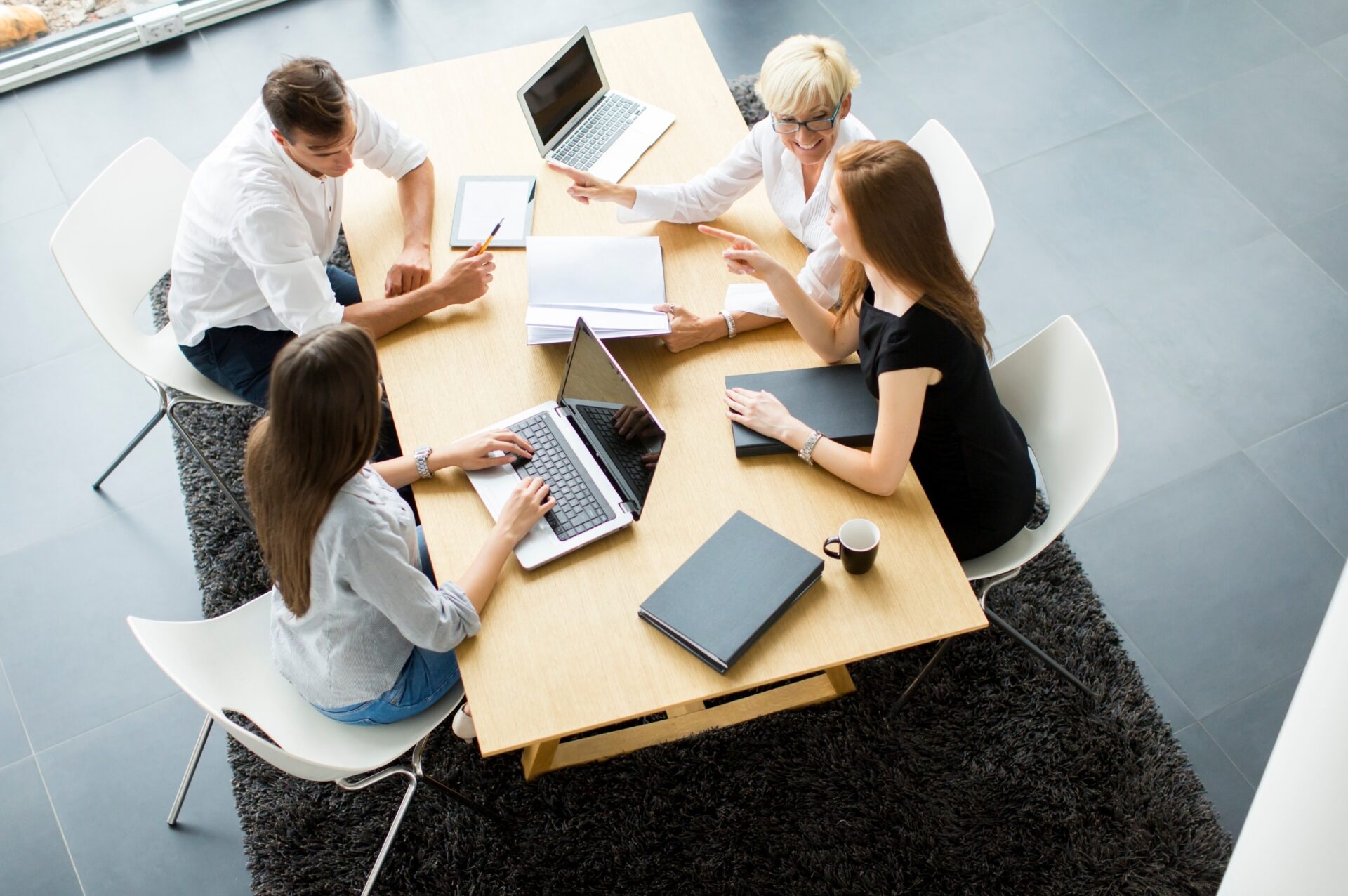 Bovenaanzicht van vier collega’s die rond een houten tafel in een moderne kantoorruimte overleg voeren met laptops, notitieboeken en een koffiekopje.