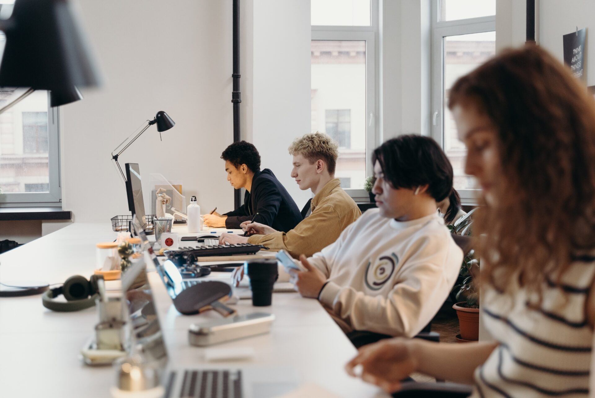 Vier mensen werken geconcentreerd aan een lange tafel in een lichte open kantoorruimte, met laptops, toetsenborden en bureaulampen bij grote ramen.