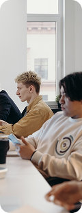 Twee mensen werken aan een tafel in een lichte vergader- of studieruimte; één typt op een laptop terwijl de ander op een smartphone kijkt, met kopjes en een raam op de achtergrond.