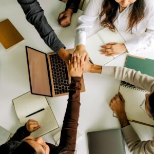 Bovenaanzicht van meerdere teamleden die hun handen op elkaar leggen boven een kantoortafel met laptop en notitieblokken als teken van samenwerking.