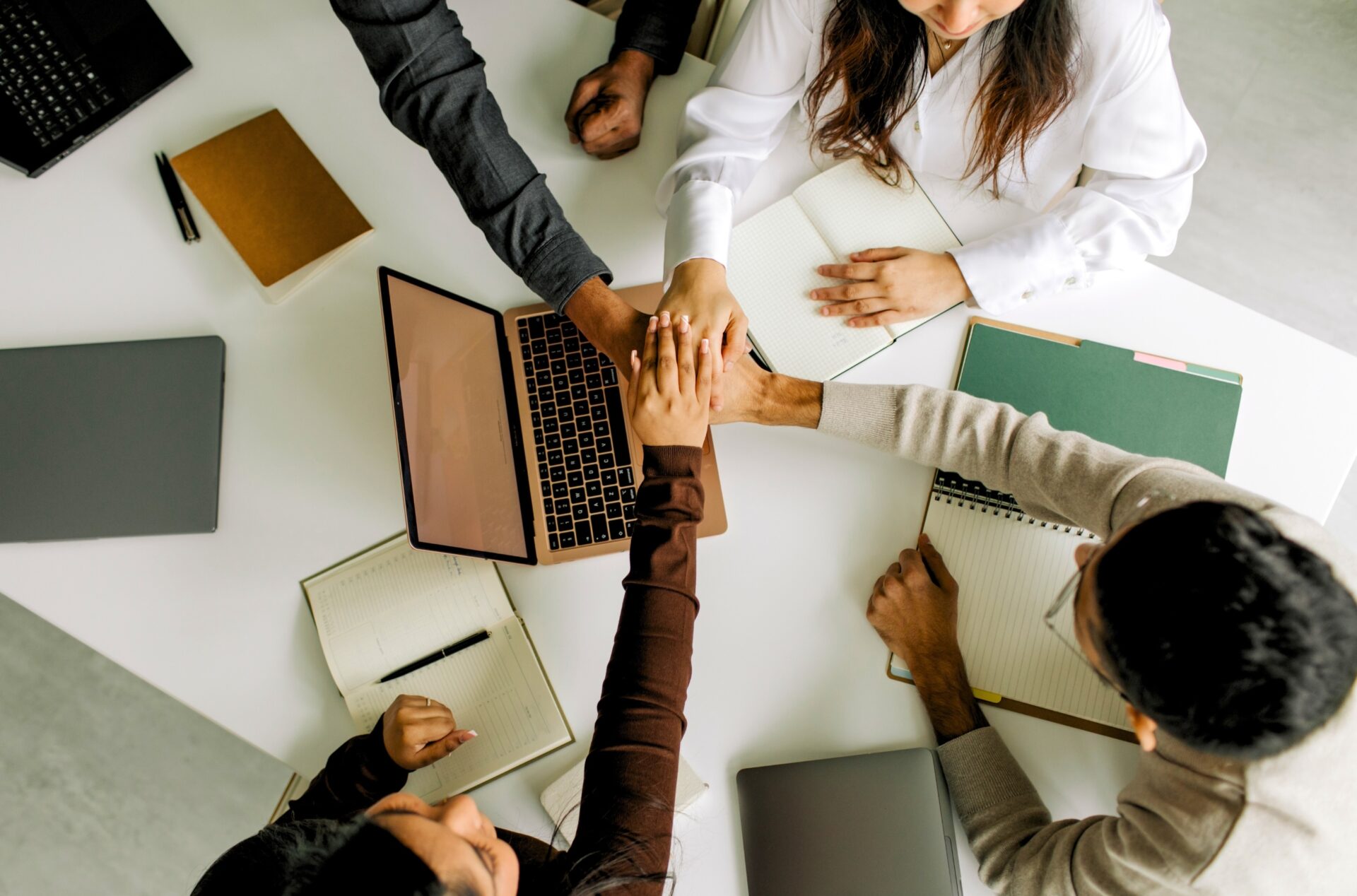 Bovenaanzicht van meerdere teamleden die hun handen op elkaar leggen boven een kantoortafel met laptop en notitieblokken als teken van samenwerking.