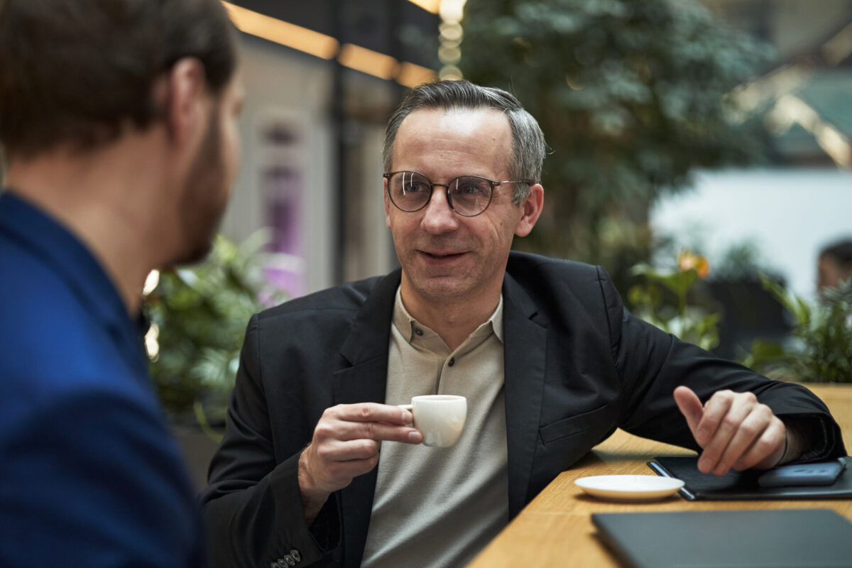 Een man met bril in een donker colbert houdt een espressokopje vast en praat met een andere persoon aan een tafel in een lichte, groene binnenruimte; op tafel liggen een schoteltje en een tablet.