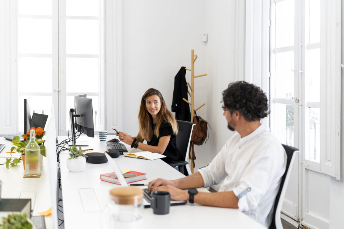 Twee collega’s praten en werken tegenover elkaar aan een licht, modern bureau met computers, notitieboek en planten in een kantoor met grote ramen.