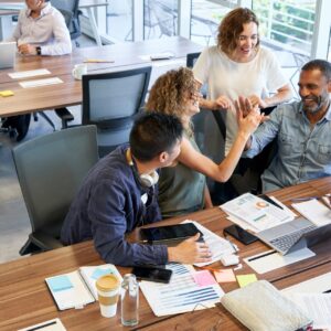 Een groep collega’s lacht en geeft elkaar een high-five rond een vergadertafel vol laptops, notities en grafieken in een moderne kantoorruimte met grote ramen.
