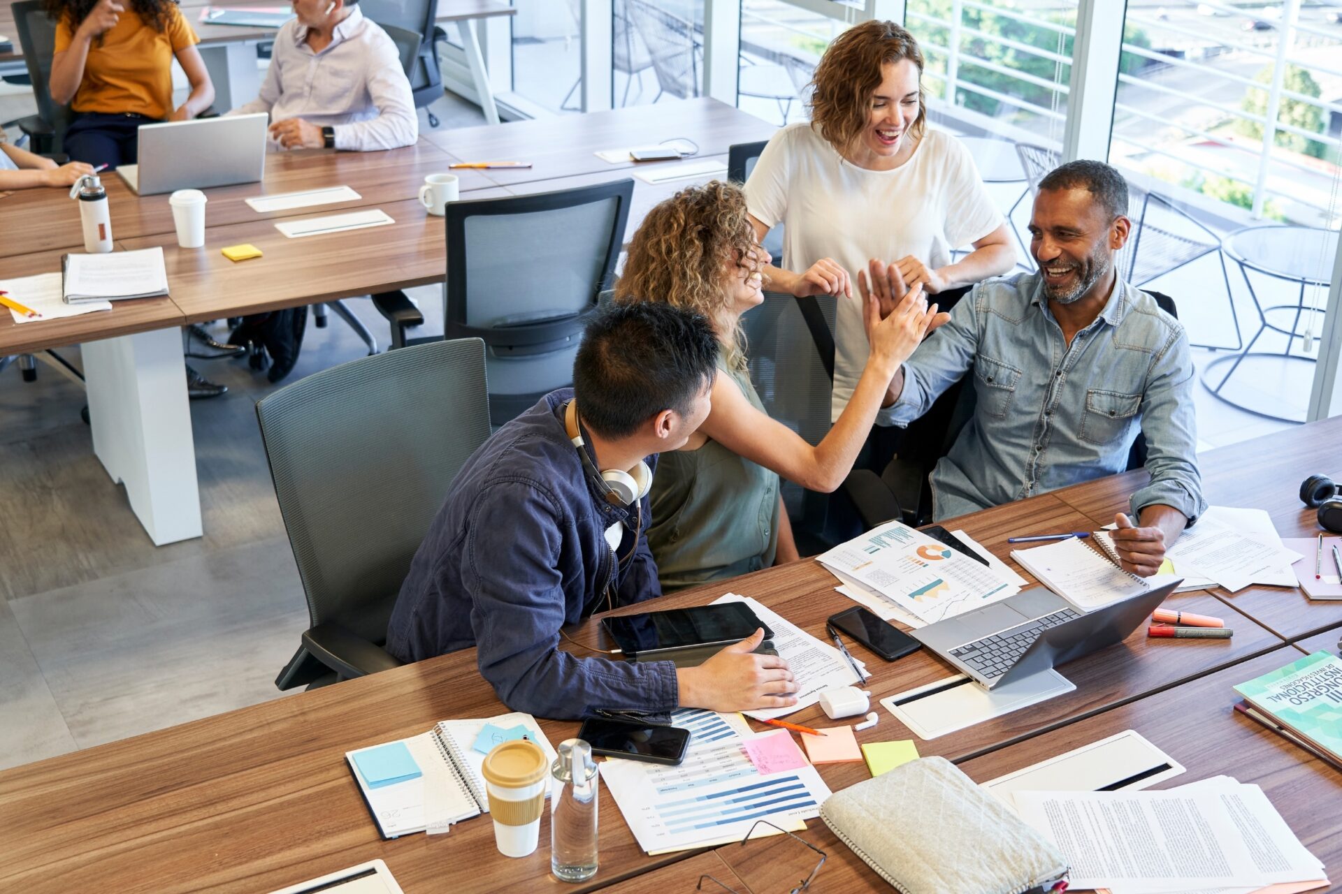 Een groep collega’s lacht en geeft elkaar een high-five rond een vergadertafel vol laptops, notities en grafieken in een moderne kantoorruimte met grote ramen.