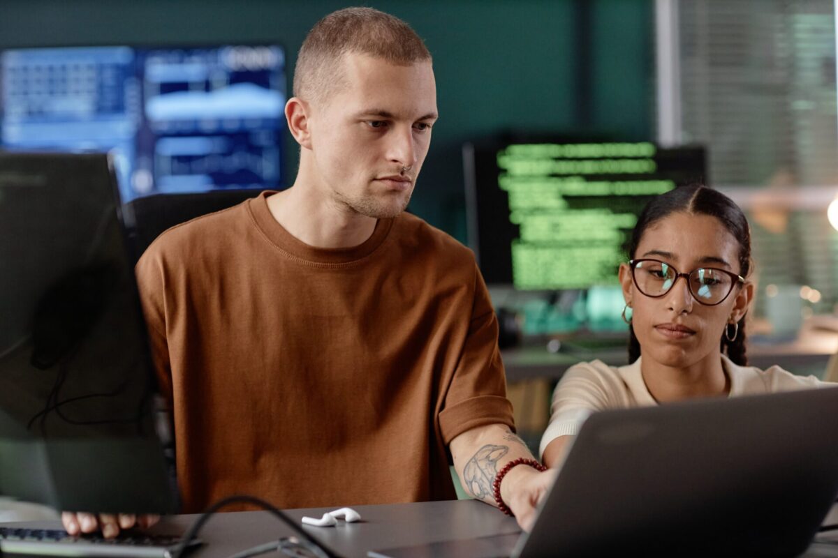 Twee collega’s werken geconcentreerd samen aan een laptopscherm, met op de achtergrond monitoren waarop code zichtbaar is.