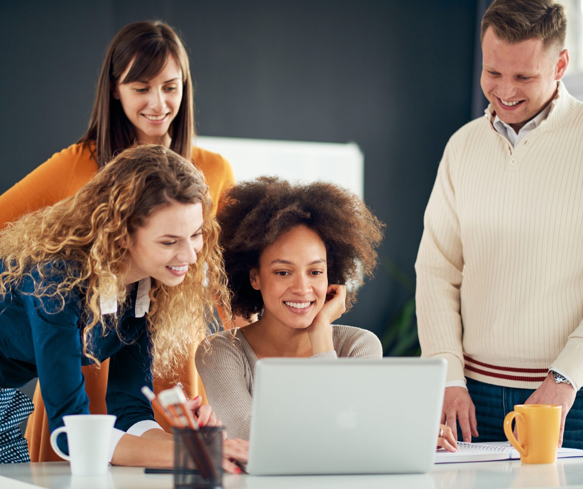 Vier collega’s kijken lachend samen naar een laptop tijdens een informele bespreking in een modern kantoor, met notitieboek en koffiemokken op het bureau.
