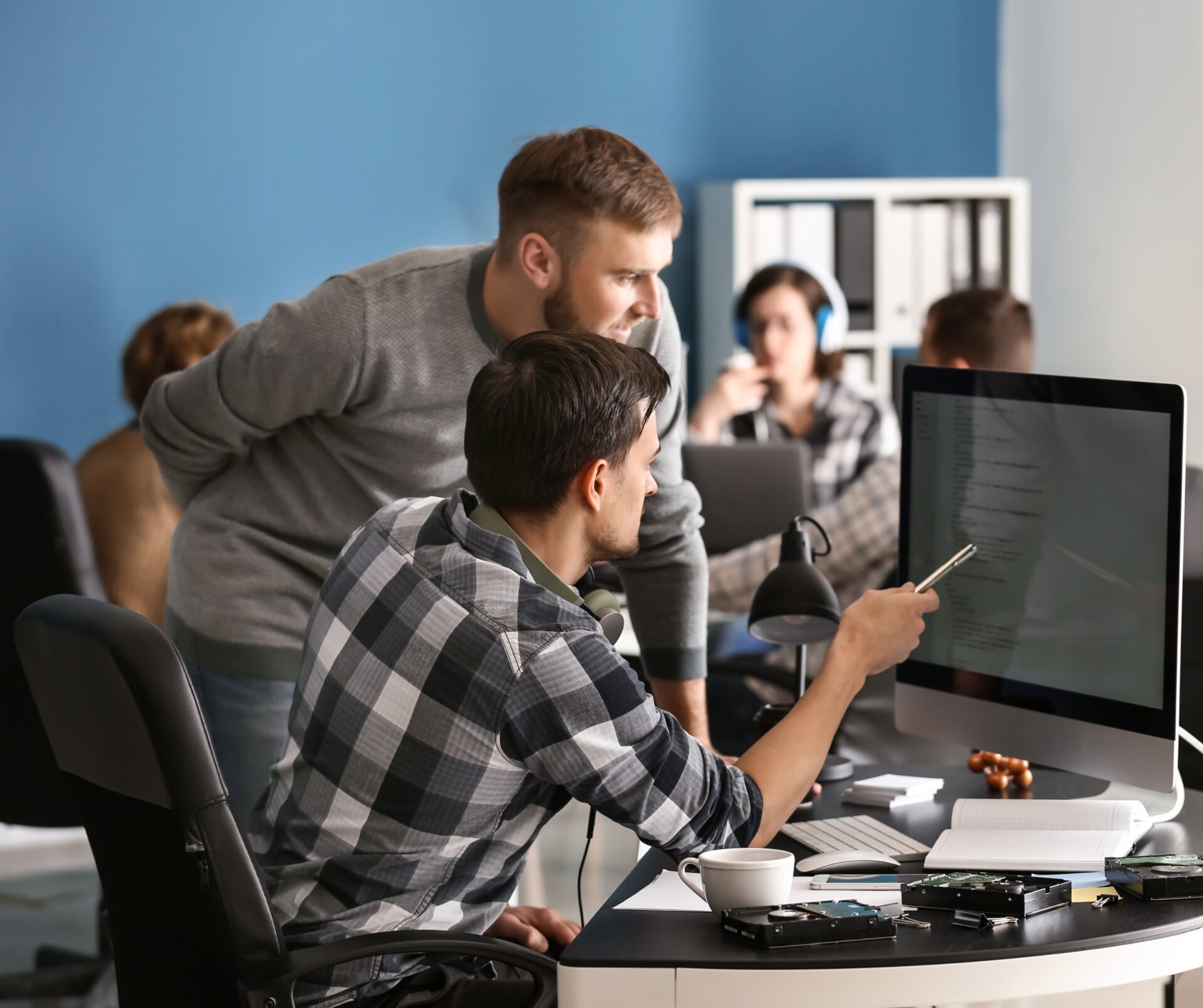 Twee collega’s in een open kantoor werken samen aan een desktop; één wijst met een pen naar code op het scherm, terwijl anderen op de achtergrond aan laptops werken.