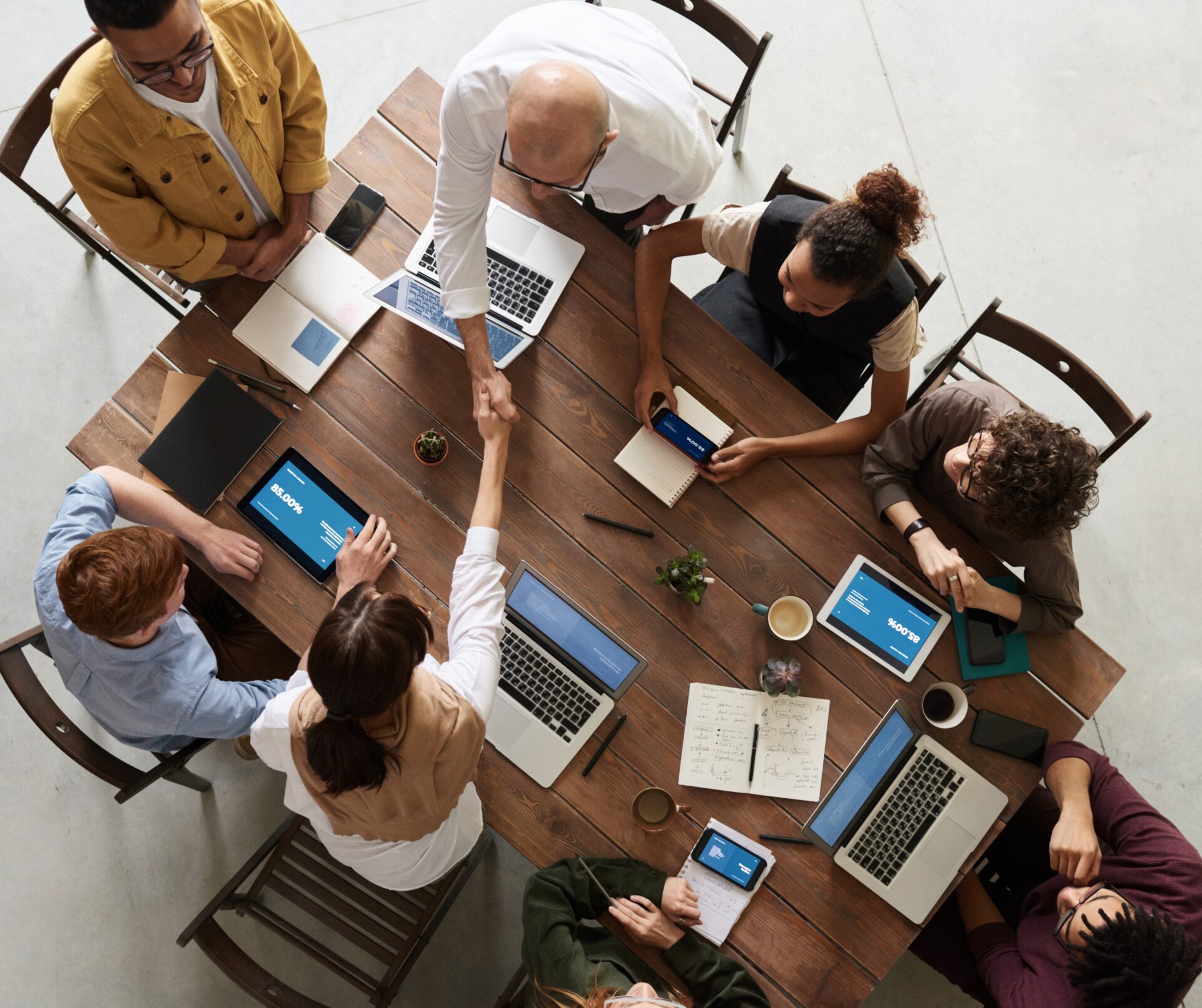 Bovenaanzicht van een teamvergadering aan een houten tafel met meerdere laptops en tablets, terwijl twee mensen in het midden elkaar de hand schudden.