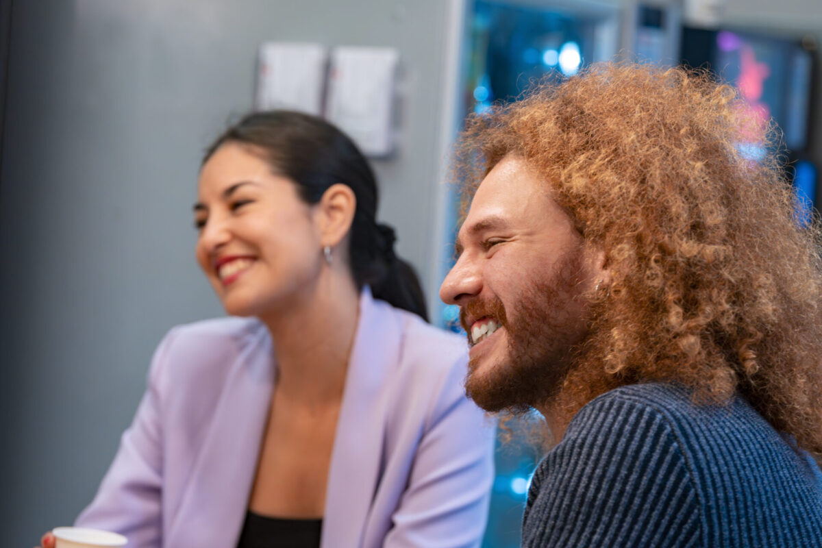 Twee lachende personen in gesprek in een informele kantooromgeving; rechts iemand met krullend rood haar en baard in een donkere trui, links iemand met donker haar in een staart en een lila blazer.