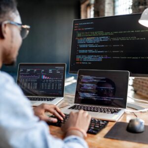 Programmeur werkt aan een bureau met twee laptops en een groot beeldscherm; op de schermen staan code en dashboards met grafieken in een moderne kantoorruimte.