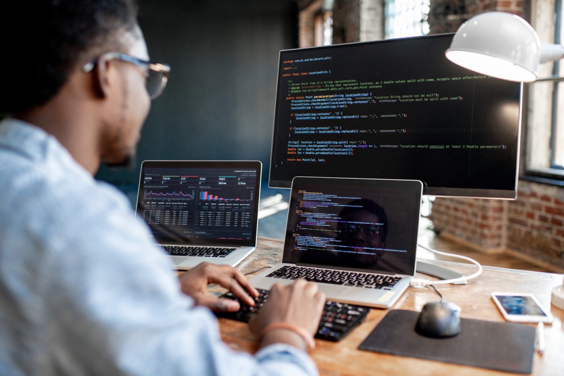 Programmeur werkt aan een bureau met twee laptops en een groot beeldscherm; op de schermen staan code en dashboards met grafieken in een moderne kantoorruimte.