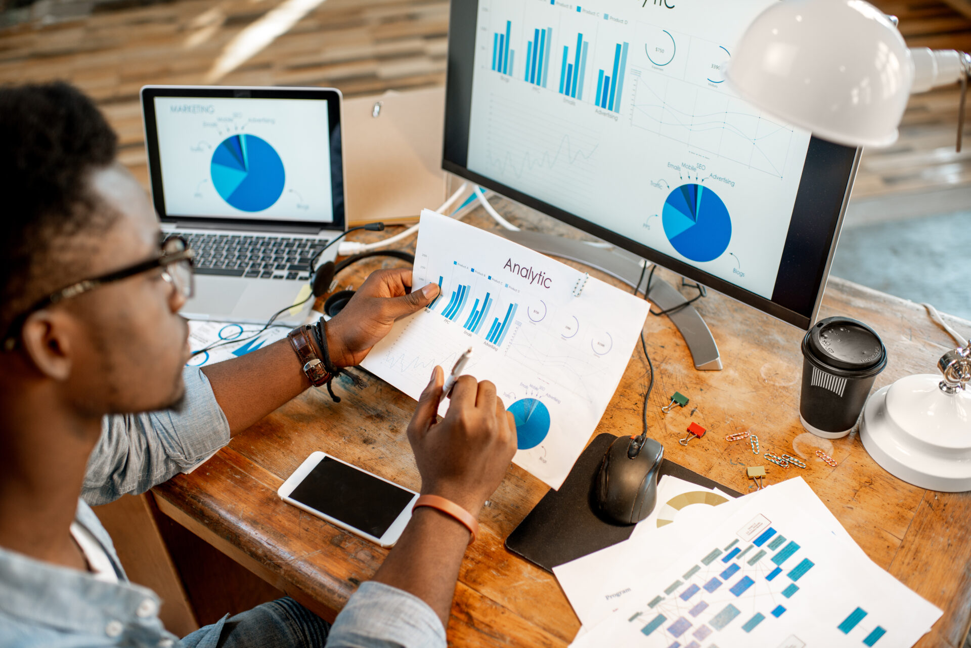 Persoon aan een houten bureau bekijkt een geprinte pagina met grafieken terwijl op laptop en monitor bar- en cirkeldiagrammen met data-analyse staan, omringd door smartphone, muis en koffiebeker.