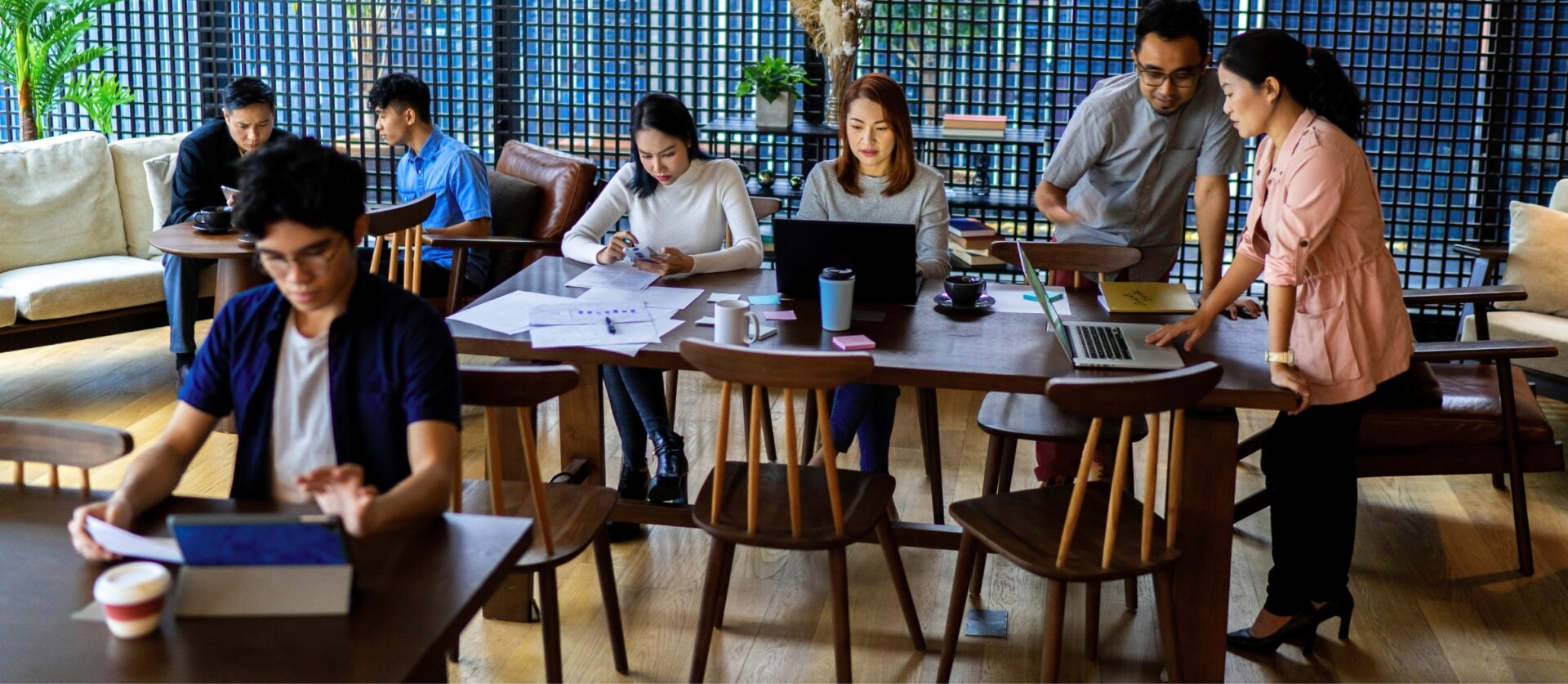 Een groep jonge professionals werkt samen aan een lange houten tafel in een moderne coworkingruimte, met laptops, papieren en koffie, terwijl anderen op de achtergrond overleggen.