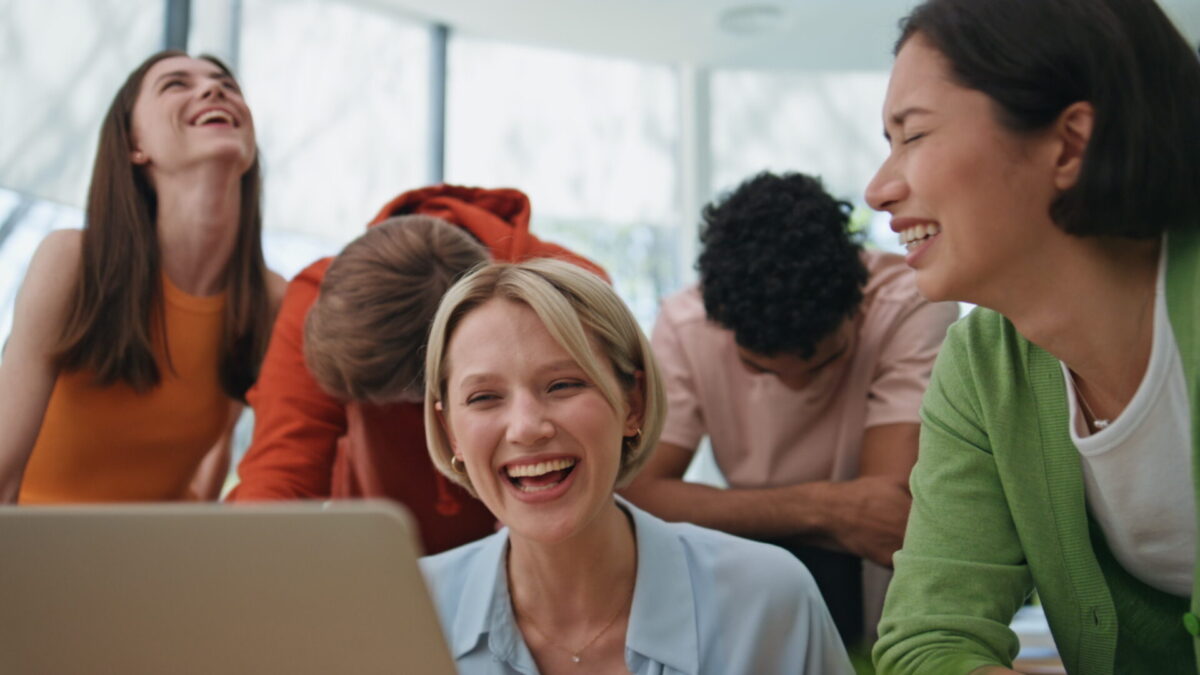 Vijf mensen lachen samen rond een laptop in een lichte ruimte met grote ramen, met de vrouw in het midden als focuspunt.