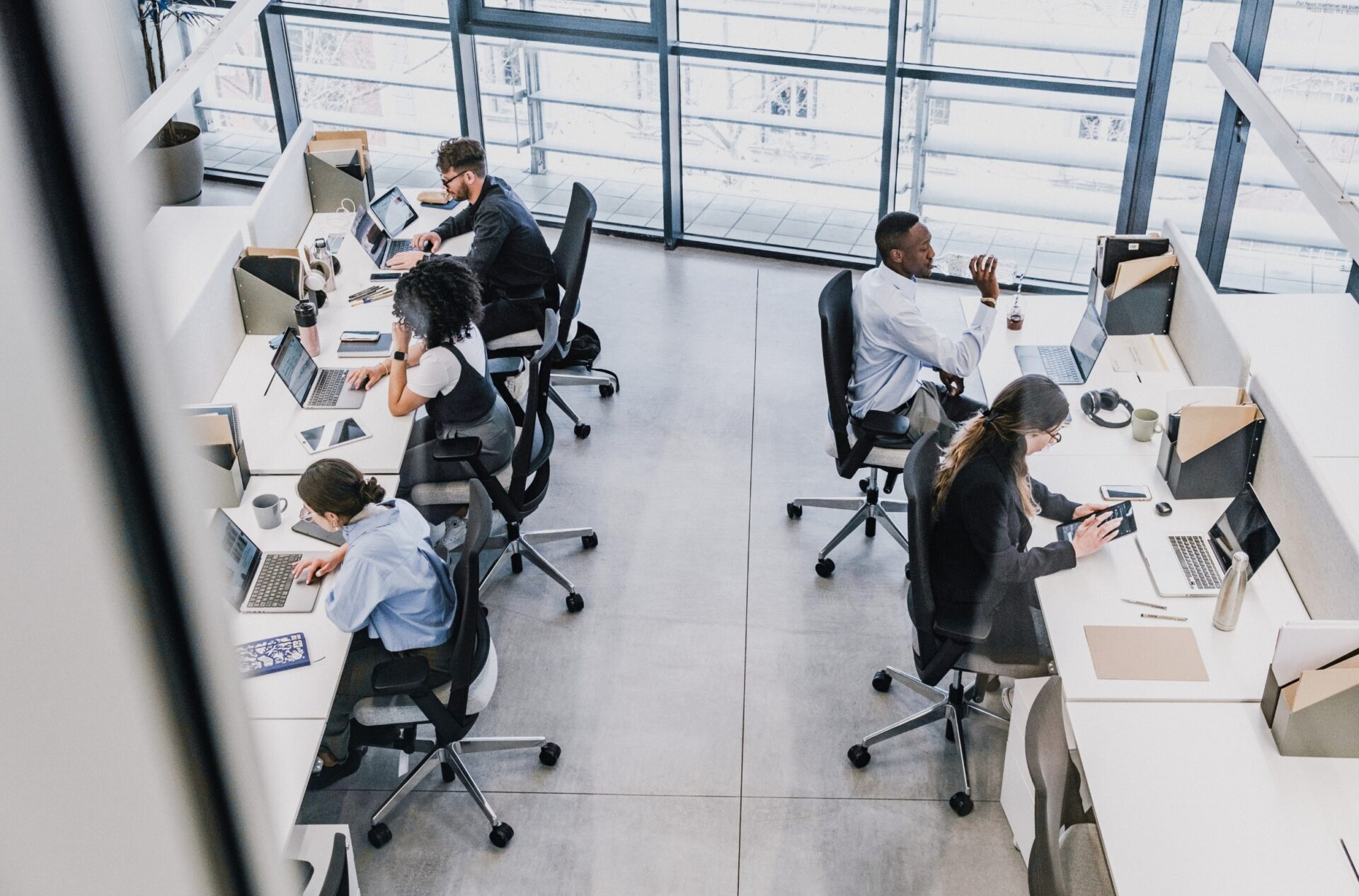 Lichte open kantoorruimte met vijf mensen die aan tegenoverliggende bureaus op laptops werken, gezien van boven, met grote ramen en moderne kantoorspullen zoals koptelefoons, notitieblokken en koffiemokken.