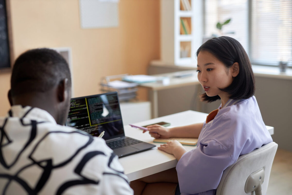 Twee collega’s aan een bureau bespreken code op een laptop, terwijl één met een pen naar het scherm wijst in een lichte kantoorruimte.
